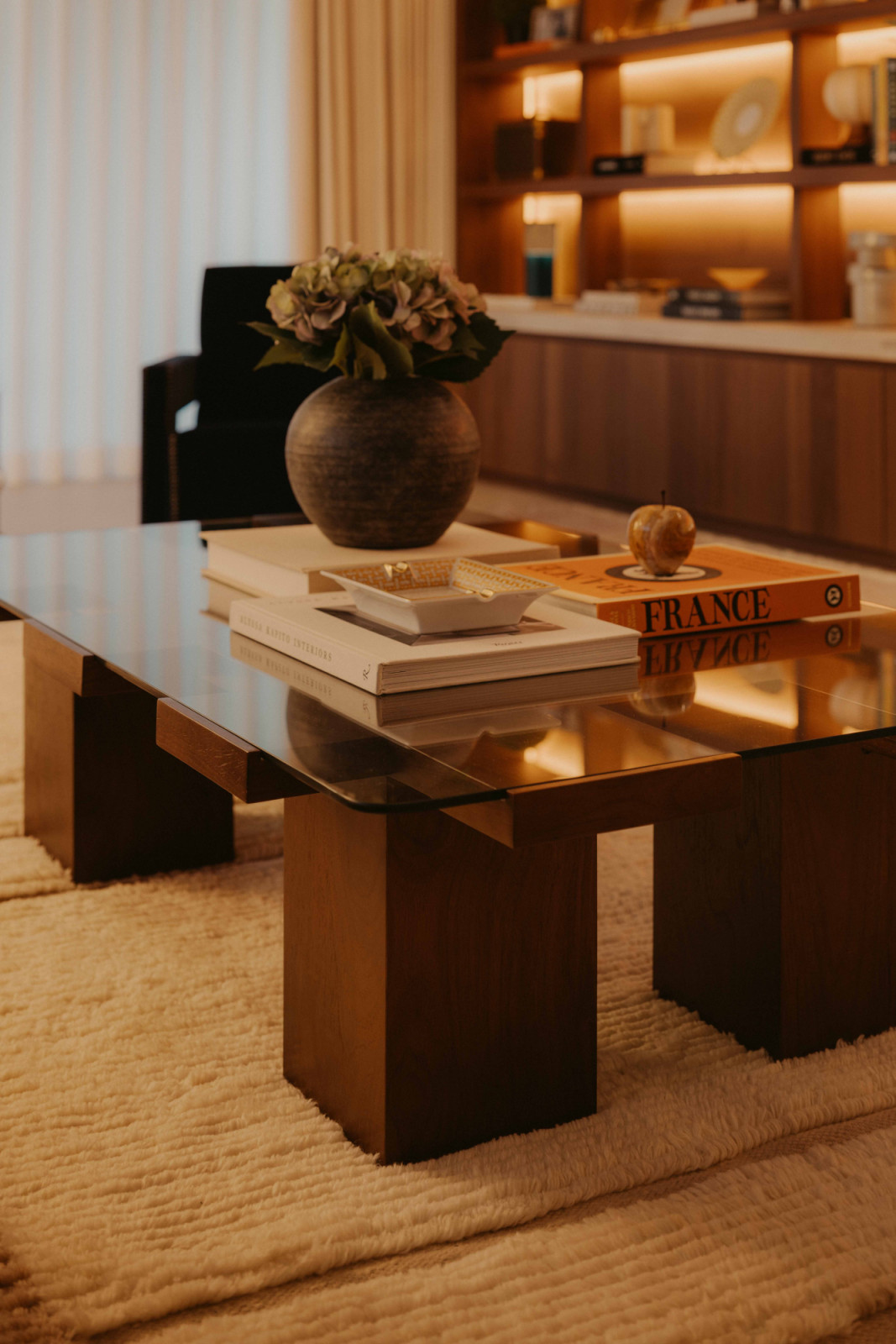 In the living area of this West Coast home, a low glass coffee table sits on a textured rug, layered with books and objects against softly lit shelving