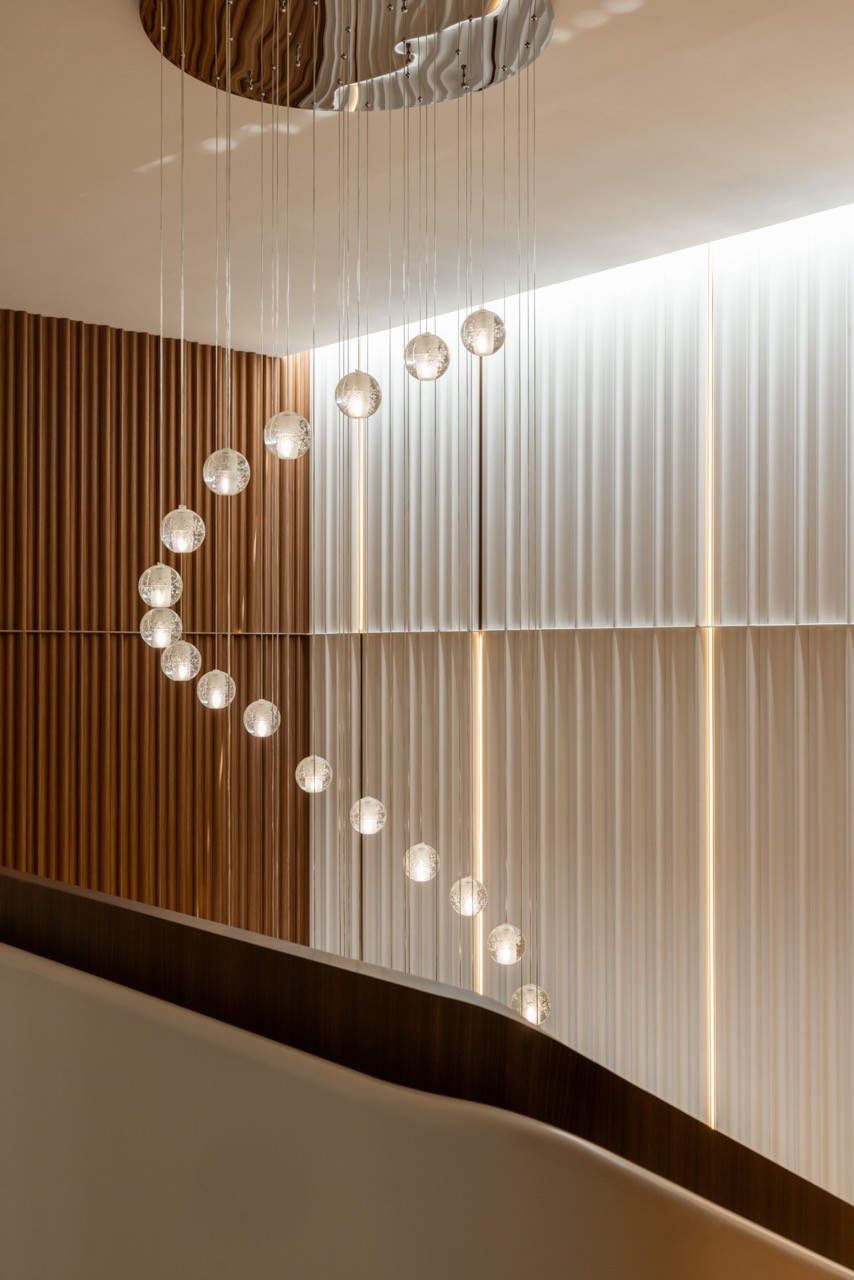 The pendant cluster from below, showing its full drop against the contrasting cream fluted and timber slat walls of the staircase volume.
Looking straight down through the bronze spiral staircase from the upper level, showing the full geometry of the stone treads winding to the