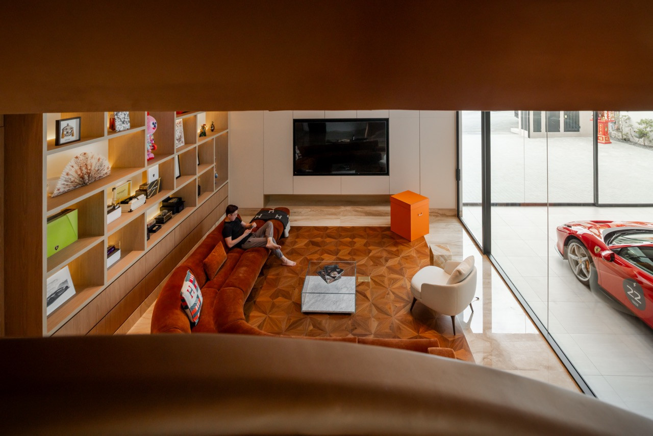 View from the spiral staircase looking down into the car lounge, with the display shelving, rust-orange velvet sofa, and parquet flooring below; a Ferrari is visible through the glass partition to the right