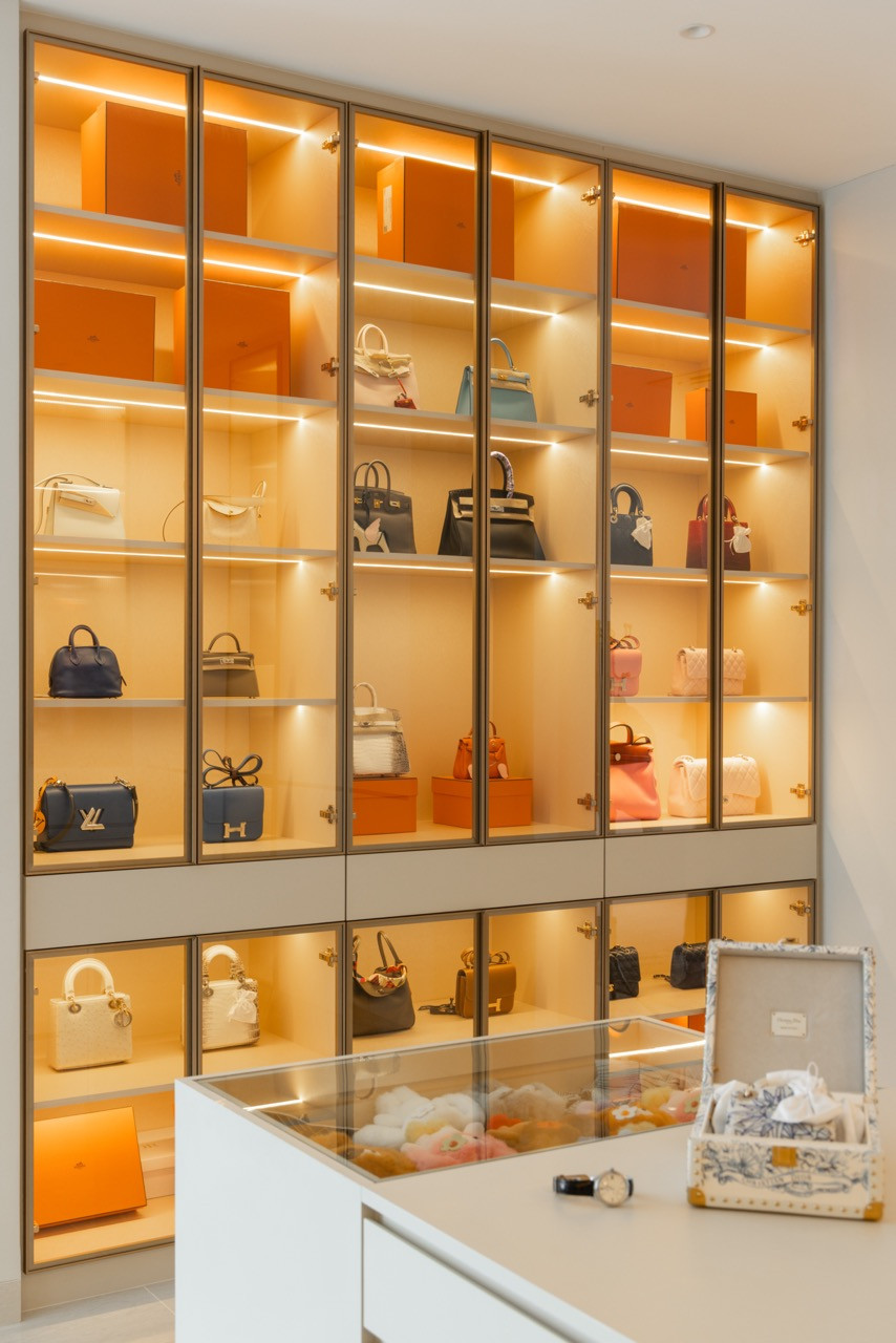 The walk-in wardrobe display cabinet, with backlit glass shelves holding a collection of handbags and a glass-topped jewellery island in the foreground