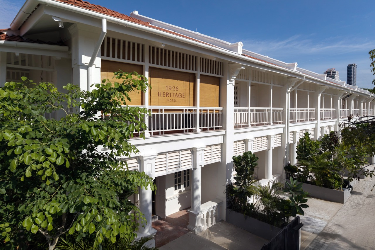 The full length of the Jalan Burma facade, with the continuous first-floor verandah and terracotta roof tiles intact; the towers of contemporary Penang visible beyond
