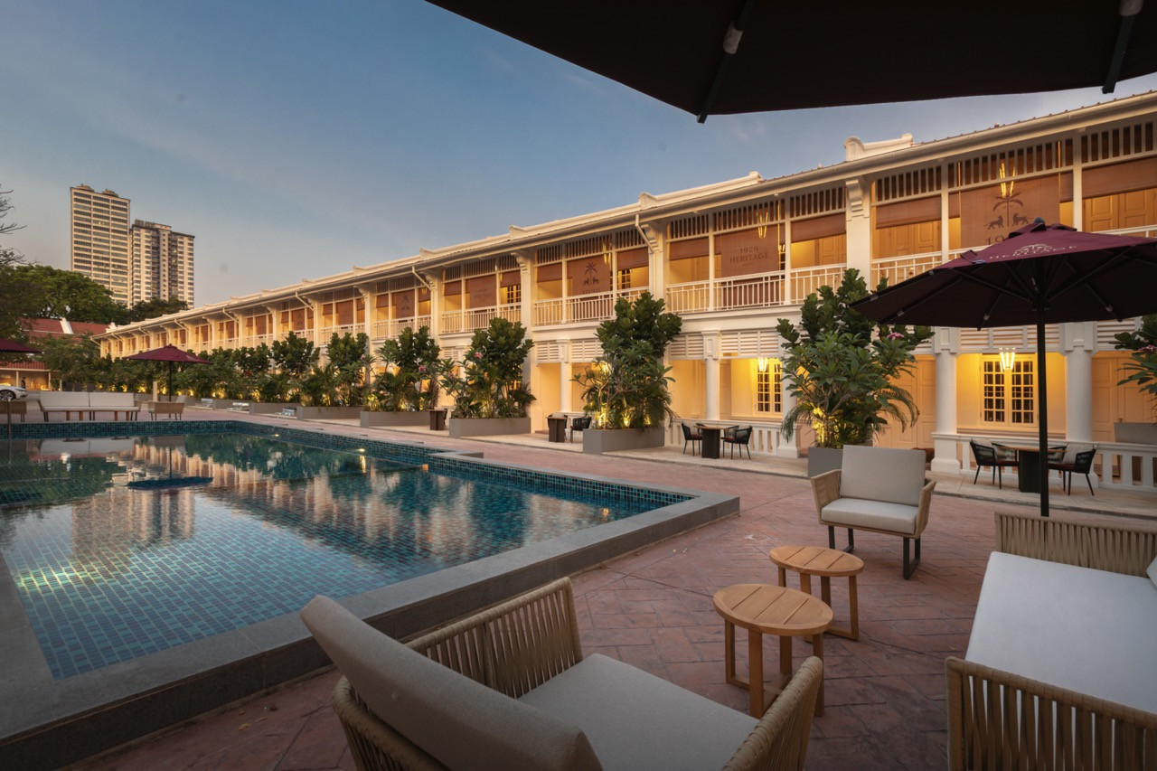 The pool terrace at dusk, with the full run of the complex visible behind; contemporary seating in the foreground and the towers of modern Penang beyond