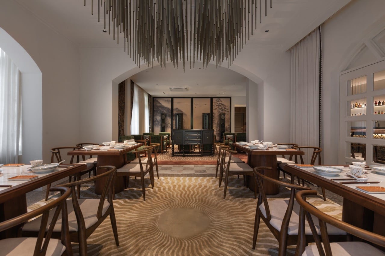 The dining room of Root House by Gēn, with dark timber wishbone chairs, marble-topped tables set with Chinese porcelain and chopsticks, and a landscape ink painting visible through the arched passage beyond