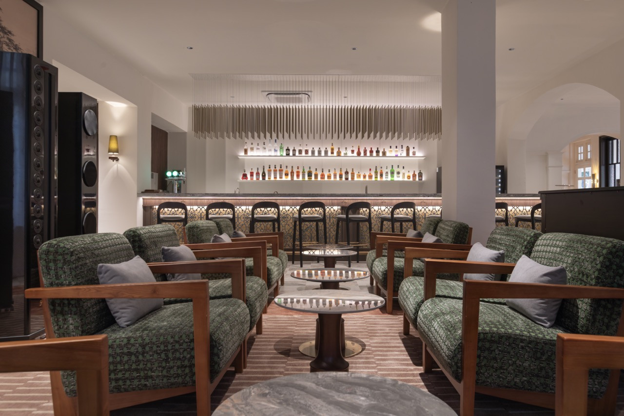 U-Bar, with green-upholstered timber armchairs grouped around low tables in the foreground and the full length of the backlit bar behind; a suspended brass rod installation runs the width of the ceiling above