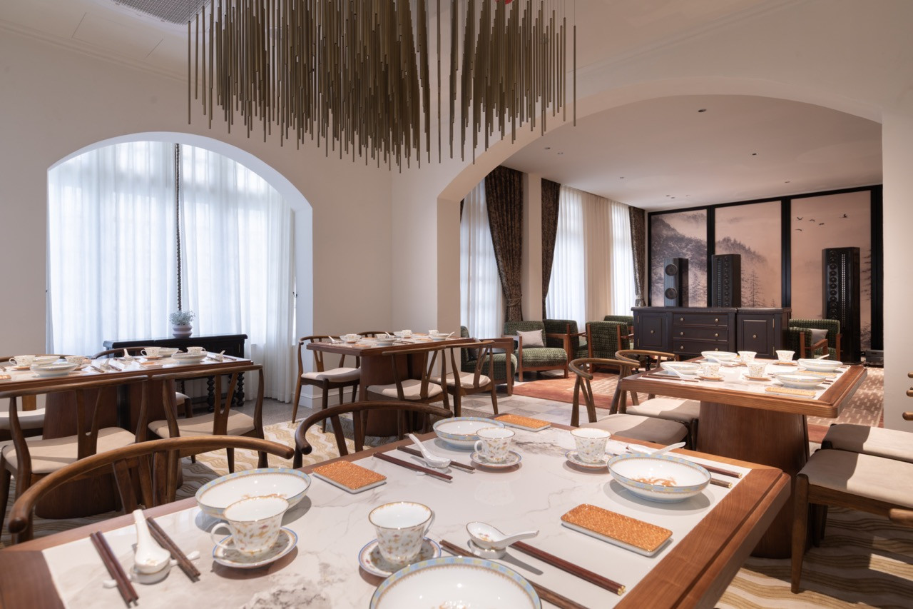 The main dining room of Root House by Gēn, with arched windows admitting natural light, dark timber furniture, Chinese porcelain place settings and the bar lounge area visible through the arch to the right