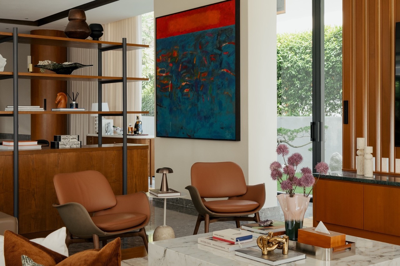 The living room features cognac leather armchairs, an open timber and steel shelving unit and a large abstract painting, with the landscaped garden visible beyond the glazed doors