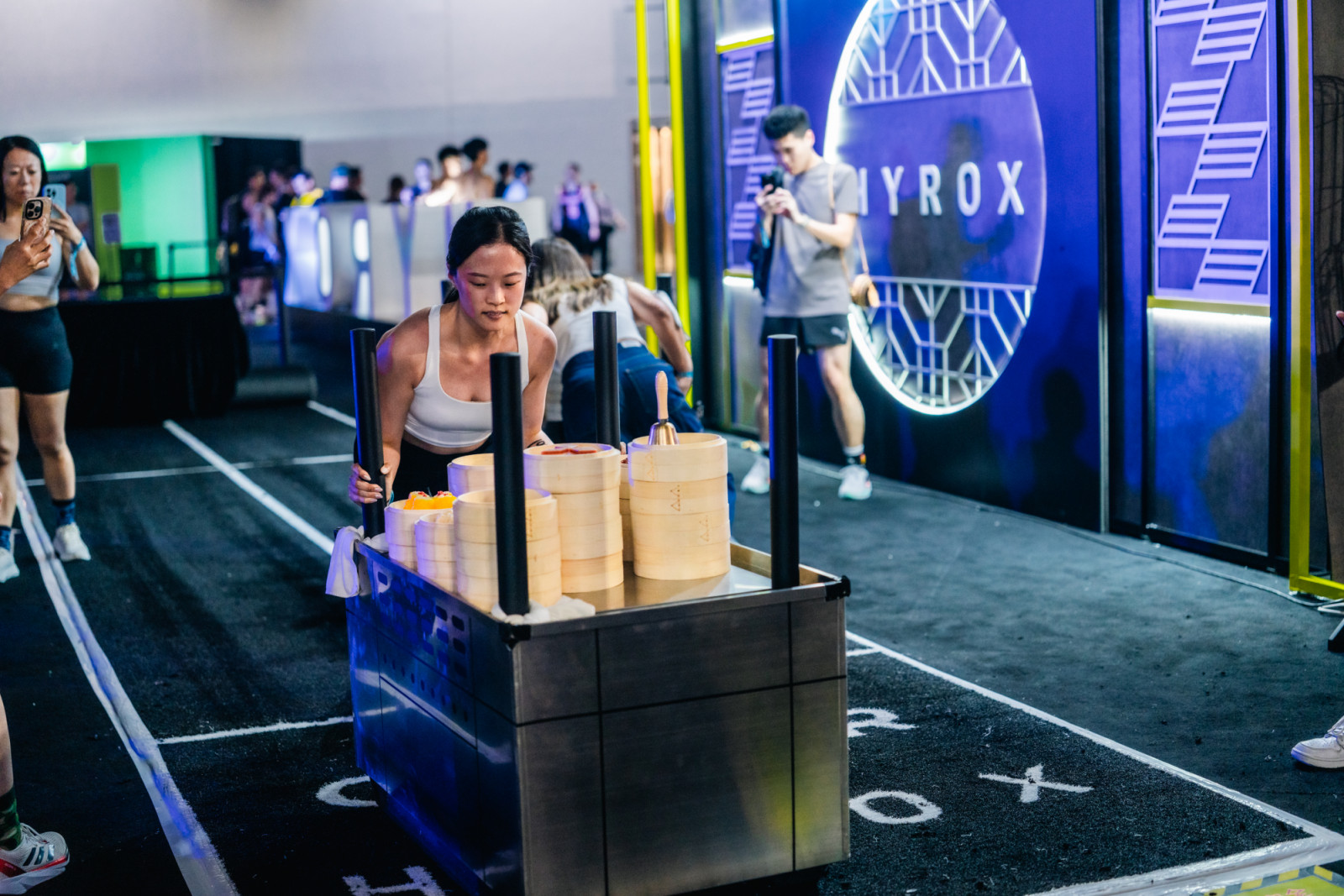 A female athlete leaning low to push a heavy metal Hyrox sled, which is uniquely loaded with stacked traditional bamboo dim sum steaming baskets