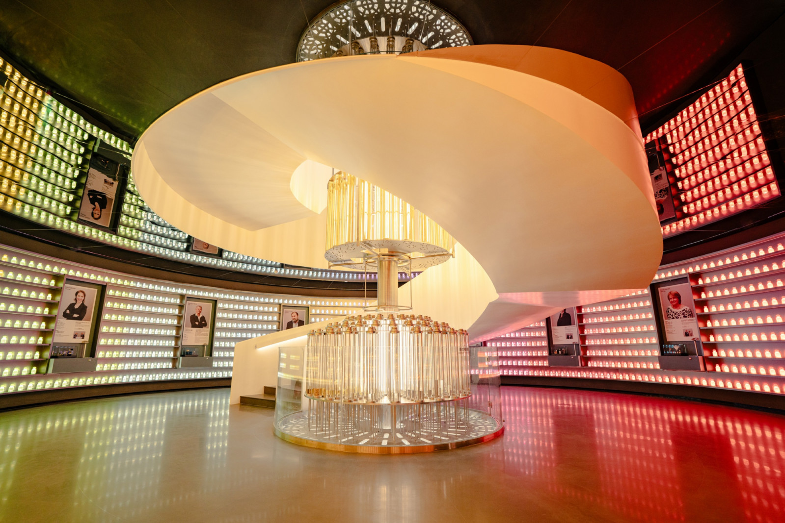 A circular gallery room lined floor-to-ceiling with illuminated fragrance vials, centred on a spiral staircase and a column of glass tubes