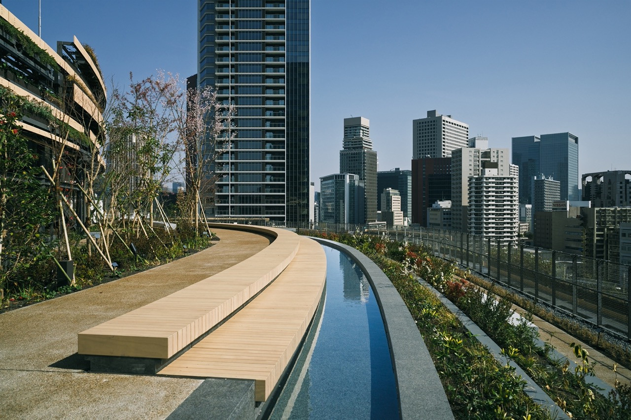 A rooftop terrace at MoN Takanawa, with a curved timber bench, a narrow reflecting pool and planted beds set against the Takanawa Gateway City tower development and the wider Tokyo skyline (Photo: Yasuyuki Takaki)