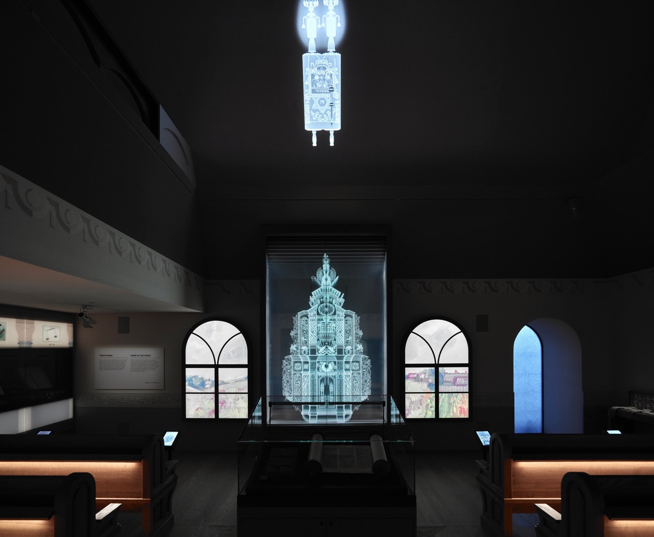 A dimly lit gallery room within the museum presents a holographic Torah scroll, with arched windows and wooden pews referencing the interior of a synagogue (Photo: Andrew Lee)
