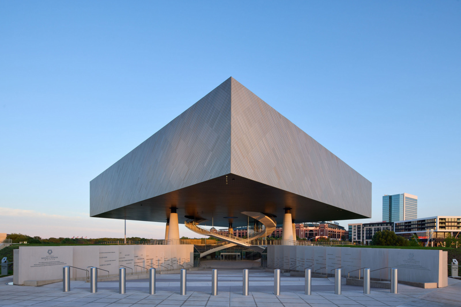 The National Medal of Honor Museum in Arlington, Texas, its steel-clad Exhibition Hall suspended 12 metres above the open-air Field of Honor courtyard on five megacolumns, with a spiral staircase visible beneath (Photo: Corey Gaffer)