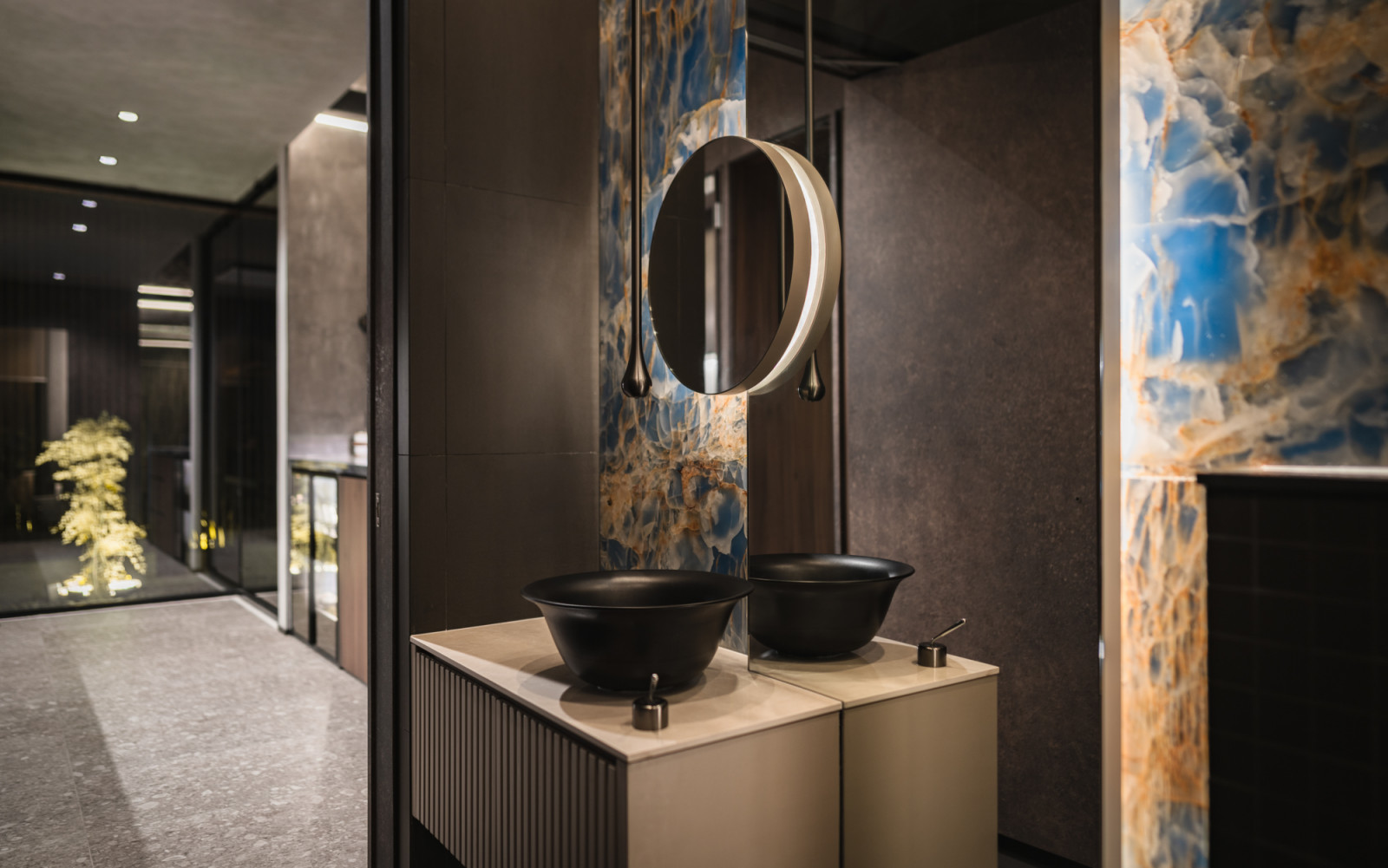 The powder room vanity with dual black matte vessel sinks, a round pendant mirror and a full-height backlit blue onyx feature wall, with a view through to the lit air well garden beyond
