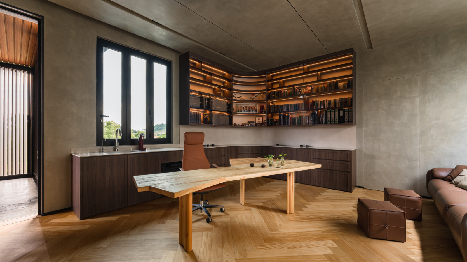 The upper floor home office showing the full-width curved corner bookshelf with amber strip lighting, the custom timber desk and leather chair, with herringbone flooring and a window facing the forested hillside