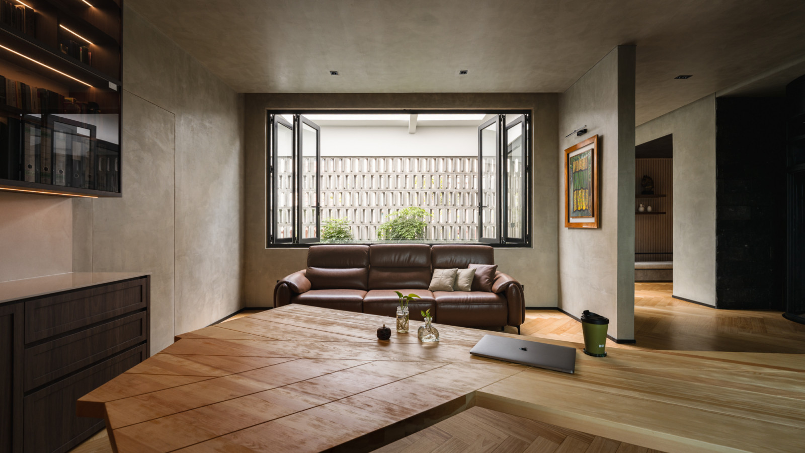 The upper floor home office with a custom timber desk of irregular profile, a curved corner bookshelf with amber lighting, herringbone timber flooring and windows on two sides overlooking a breeze block screen and garden