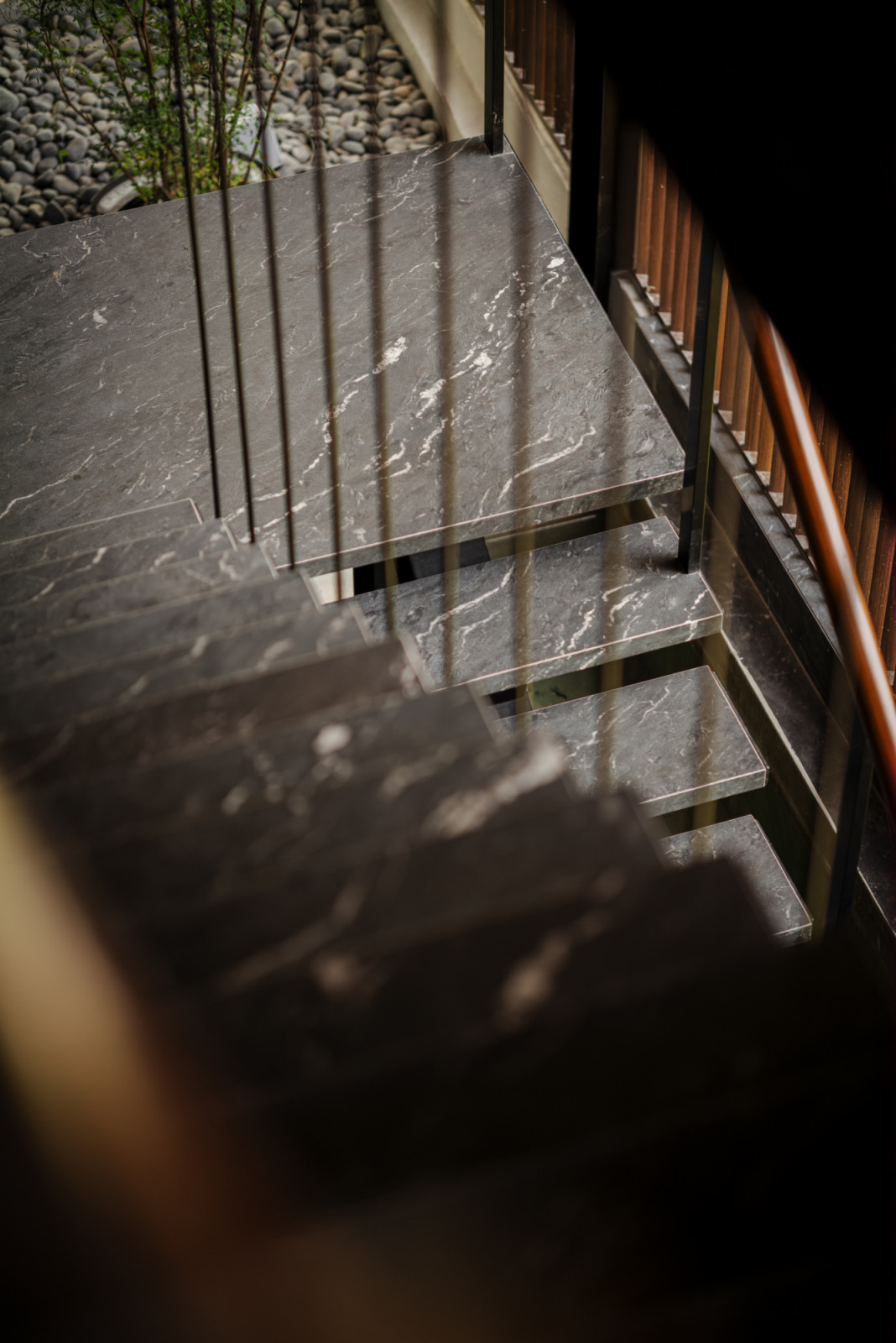 A close detail of the staircase shows dark veined stone treads cantilevered from a black steel structure, with river pebbles and planting visible through the glass balustrade below