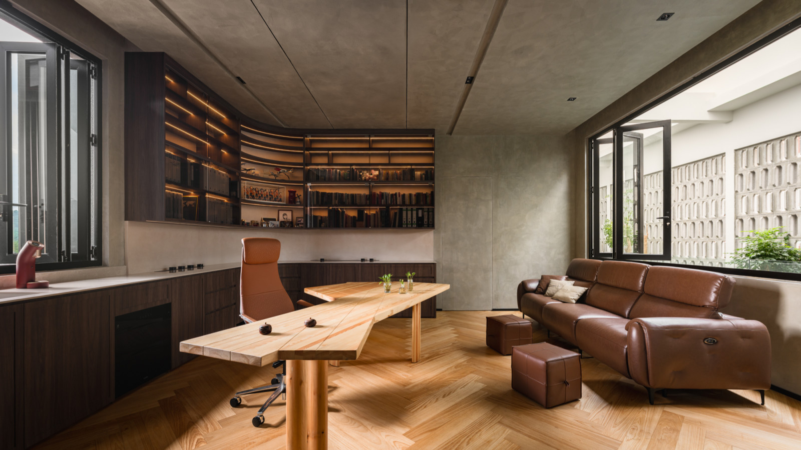 The upper floor home office from a wider angle, showing the full curved bookshelf unit, the custom timber desk, herringbone flooring, a leather sofa with ottoman to the right and a breeze block screen wall beyond the window