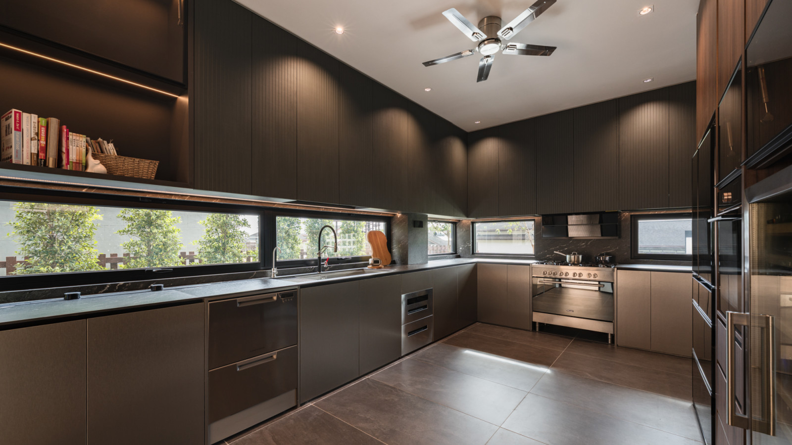 The wet kitchen looking along the work surface, showing the dark cabinetry, continuous horizontal window with garden view, stainless steel range cooker and a marble splashback panel