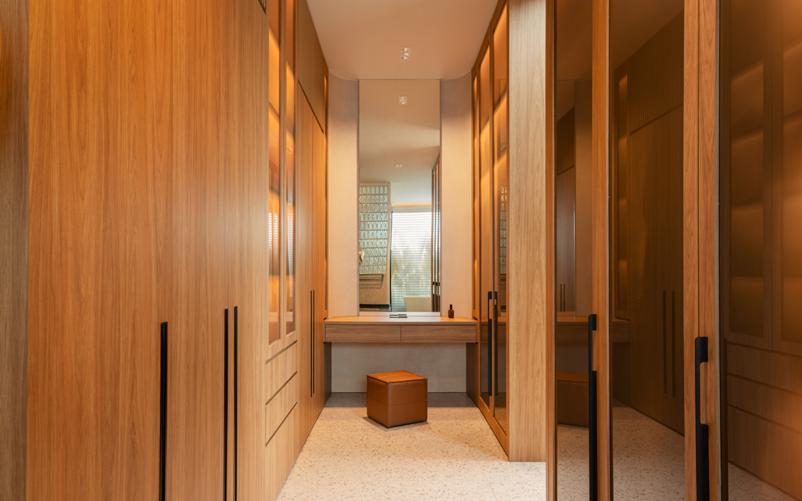 The walk-in wardrobe on the upper floor features full-height timber cabinetry with glass-fronted doors on both sides, a built-in timber dressing table with mirror at the end, and terrazzo flooring