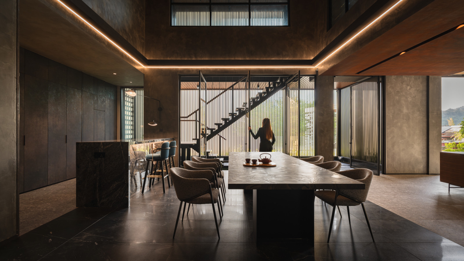 A wide view of the lower ground floor dining area showing the long stone dining table, upholstered chairs and dark marble bar island, with the open-riser staircase beyond and garden views to the right through louvred screens