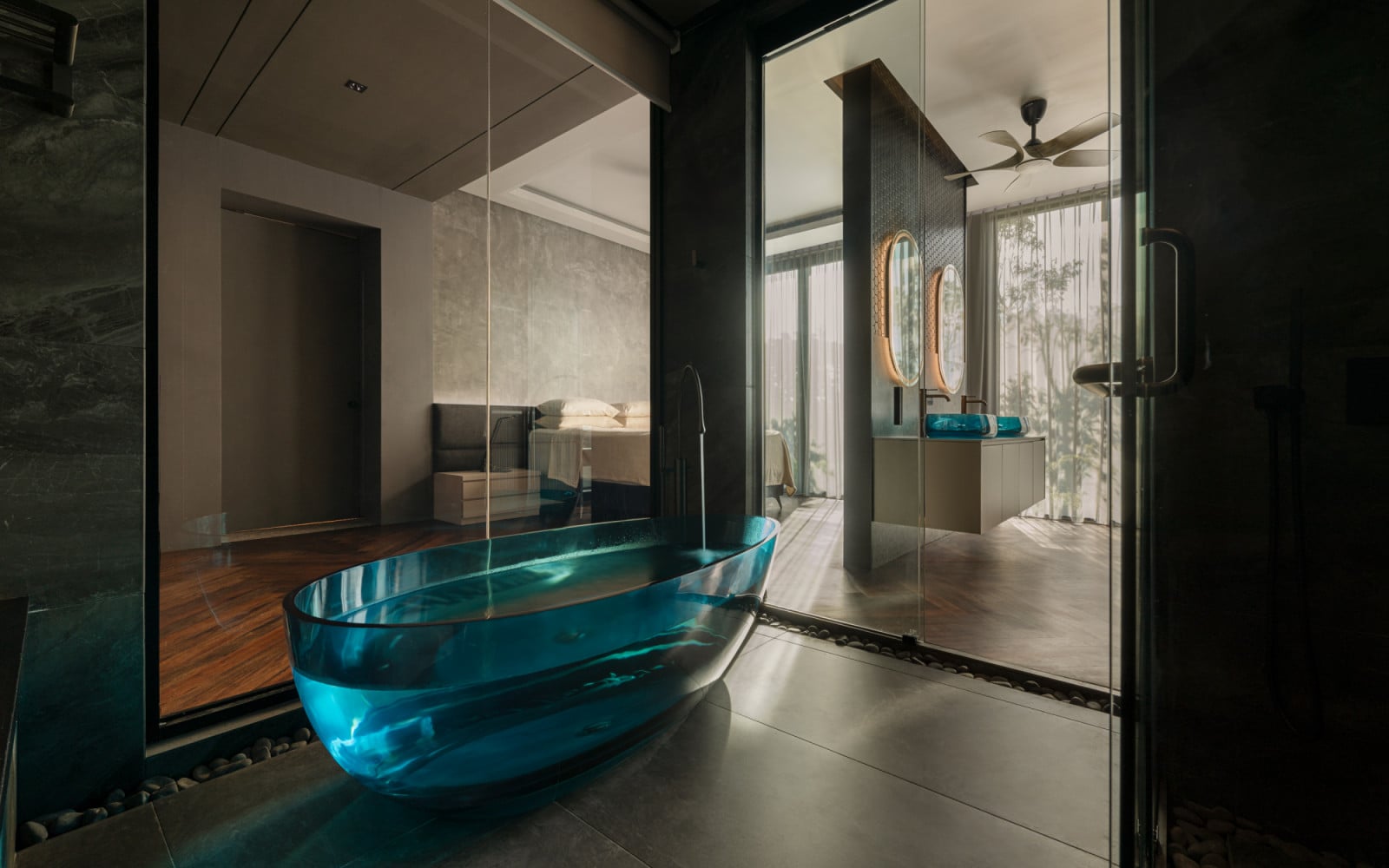 The blue glass freestanding bathtub sits between the bedroom and bathroom of the lower ground floor suite, with the dual vanity and oval backlit mirrors reflected in the glass partition behind