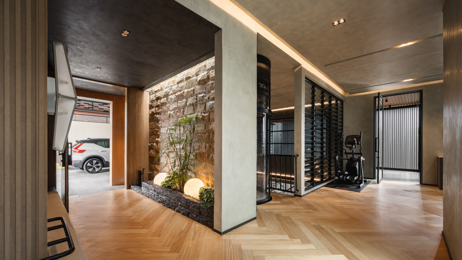 The first floor entry hall showing the stone planter niche, the panoramic elevator column, an adjacent wine display cabinet and gym equipment behind louvred screens, with herringbone timber flooring throughout