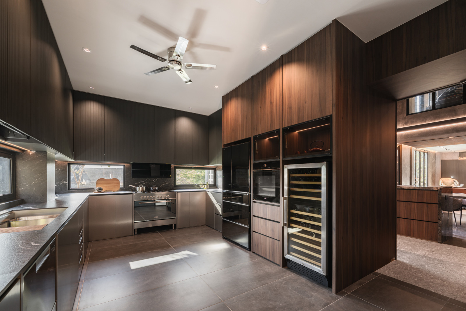 The kitchen features dark flat-front cabinetry, a stainless steel range cooker, a full-height timber-fronted appliance tower with built-in ovens and wine fridge, and a continuous horizontal window above the work surface