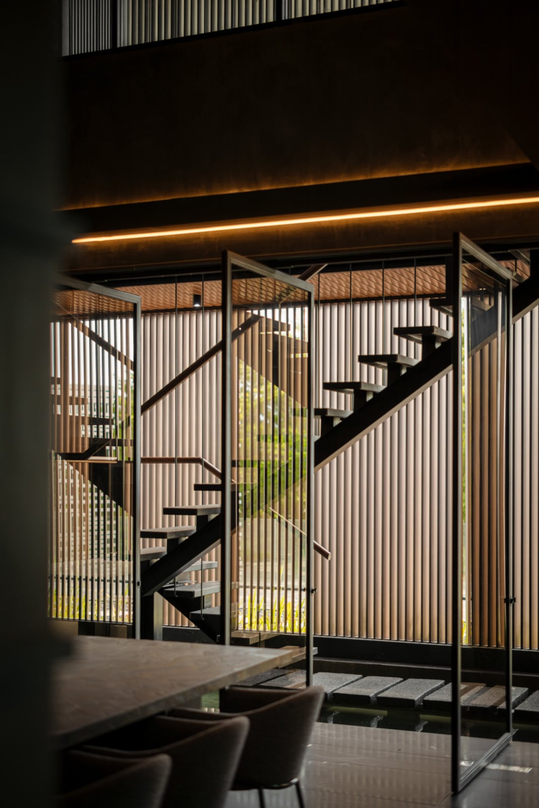 The lower ground floor dining area viewed from inside, with the open-riser staircase and vertical batten screen visible beyond the glass partition, and linear cove lighting running along the dark concrete ceiling
