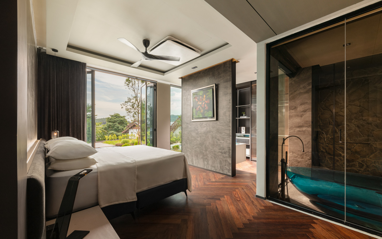 A ground floor bedroom with herringbone timber flooring, a concrete-finish headboard wall and a view through to the blue glass bathtub in the adjoining bathroom beyond the glass partition