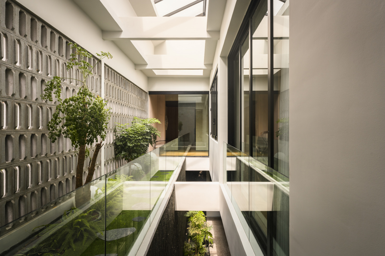The air well corridor without figures, showing the glass balustrade walkway, the planted garden bed with stepping stones and a small tree below, the breeze block screen wall and skylight above