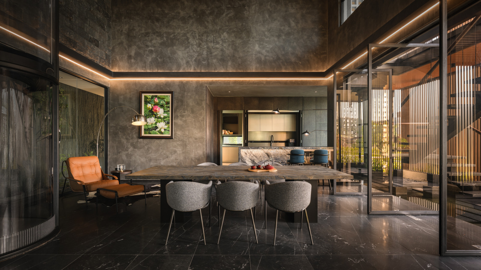A wide view of the lower ground floor dining room with the stone table and upholstered chairs at centre, the dry kitchen visible behind, a lotus painting on the raw concrete wall and the lava stone air well wall to the left