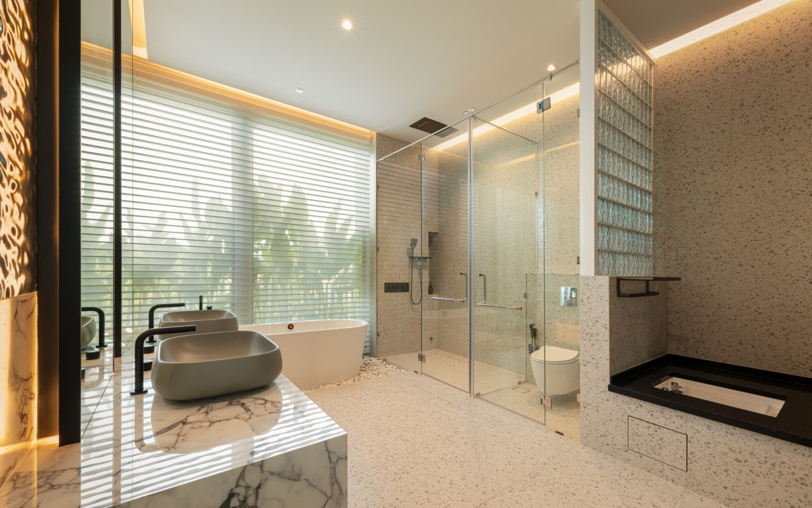 The upper floor master bathroom features dual grey vessel sinks on a marble vanity counter, a white freestanding bathtub set on river pebbles, a glass-enclosed shower and motorised horizontal blinds facing the garden