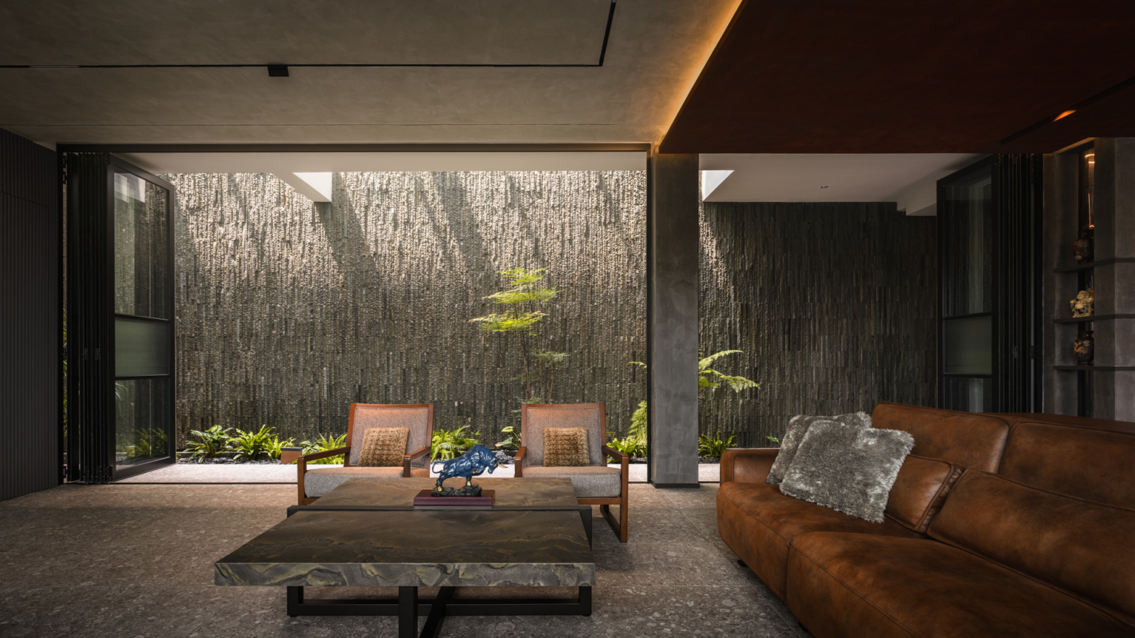 The lower ground floor living room with a tan leather sofa and timber-framed armchairs around a stone coffee table, facing a full-width textured stone feature wall with planting at its base and a skylight above