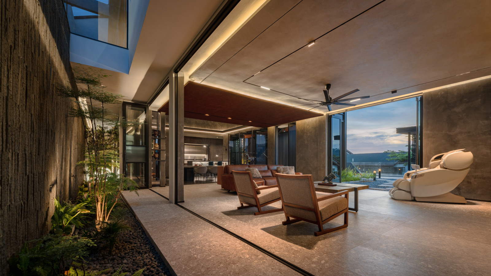 The lower ground floor living area at dusk viewed from the side, showing the lava stone air well wall to the left, the seating arrangement and the garden opening to the right through retractable glass walls