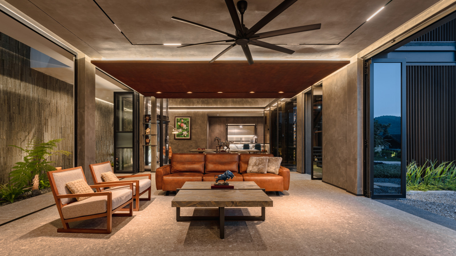 The lower ground floor living area at dusk, with a tan leather sofa and timber-framed armchairs arranged around a stone coffee table under a large ceiling fan, the dry kitchen visible in the background and garden beyond