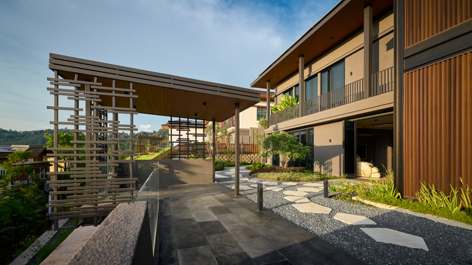 A covered walkway runs along the side of House 37, where a timber pergola structure, stone paving and gravel garden connect the building to its forested hillside setting