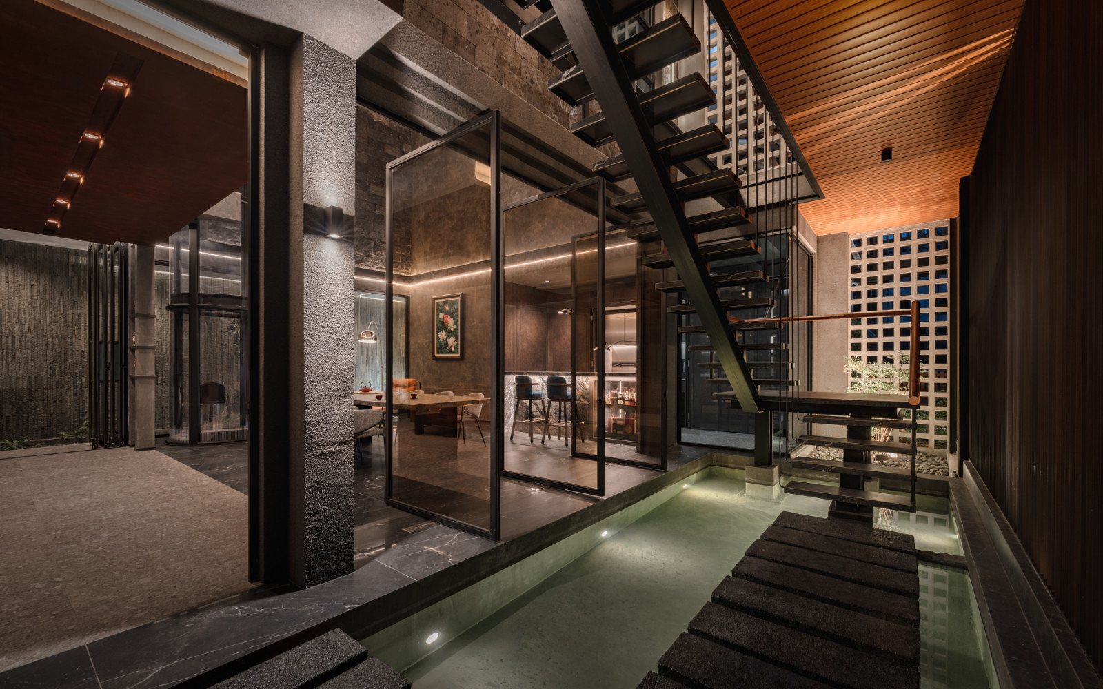 At night, the lower ground floor entry corridor shows the indoor pond lit from below, stepping stones crossing the water, the open-riser staircase, and the dining room visible through glass partitions