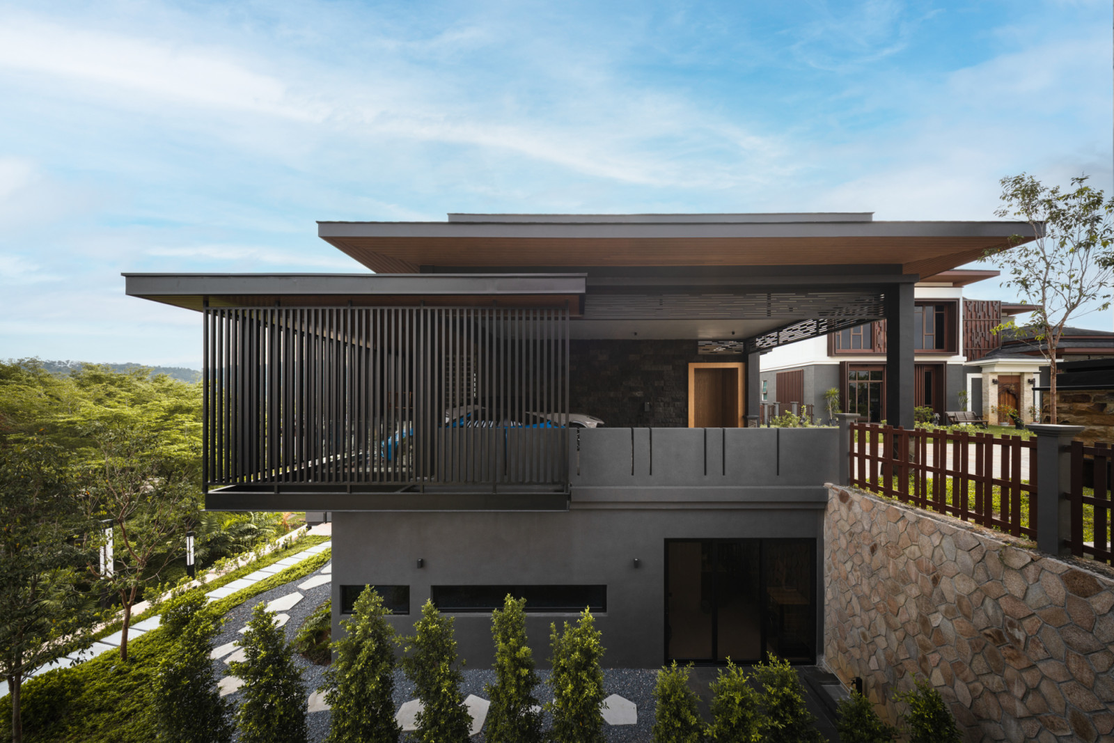 The rear elevation of House 37 showing the upper floor terrace with vertical metal batten screen, overhanging timber-lined roof, the lower ground floor with horizontal clerestory windows, a stone boundary wall and planted garden below