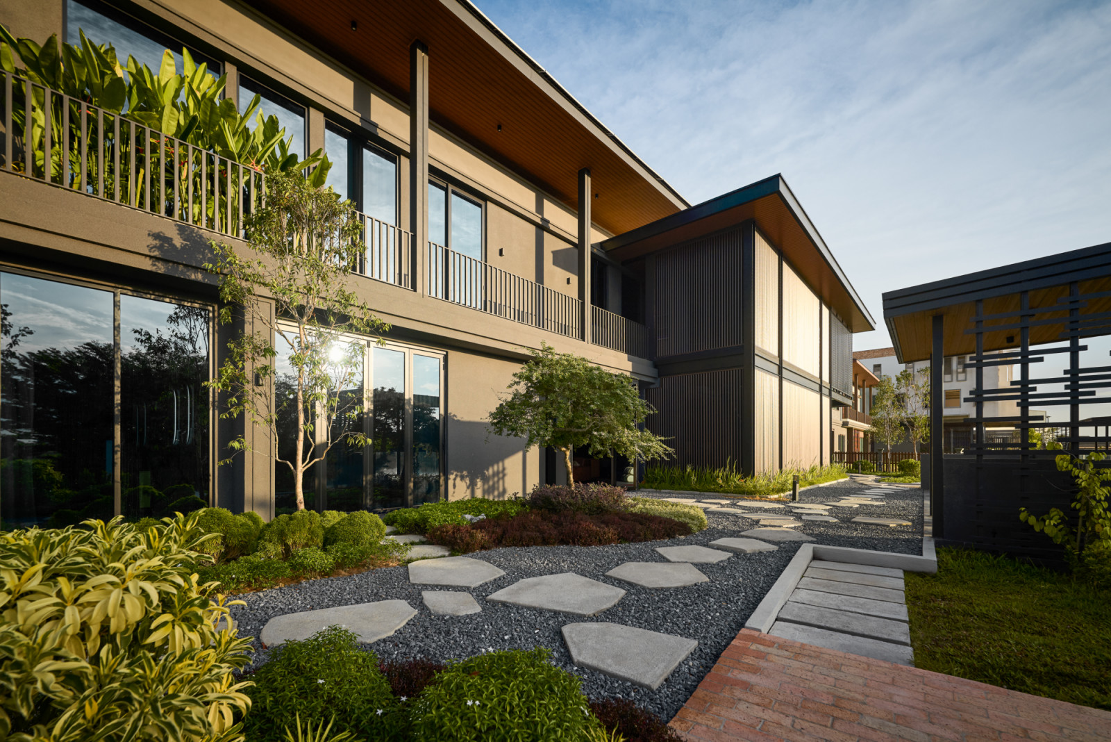 A wide view of the two-storey bungalow shows the solar-panelled roof, landscaped garden with stepping stones and a pergola structure at the garden's edge, with a forested hillside behind