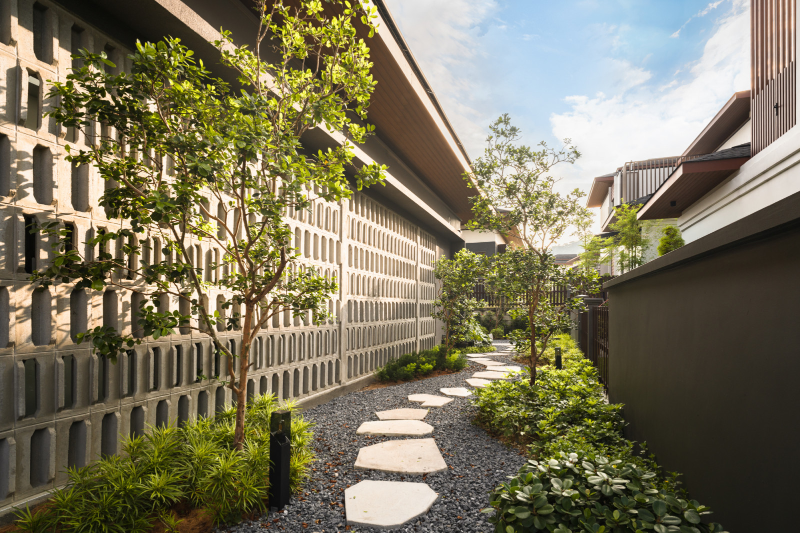 The side garden pathway with large hexagonal stone stepping stones set in dark gravel running alongside the perforated concrete breeze block screen wall, with trees and low shrubs on both sides