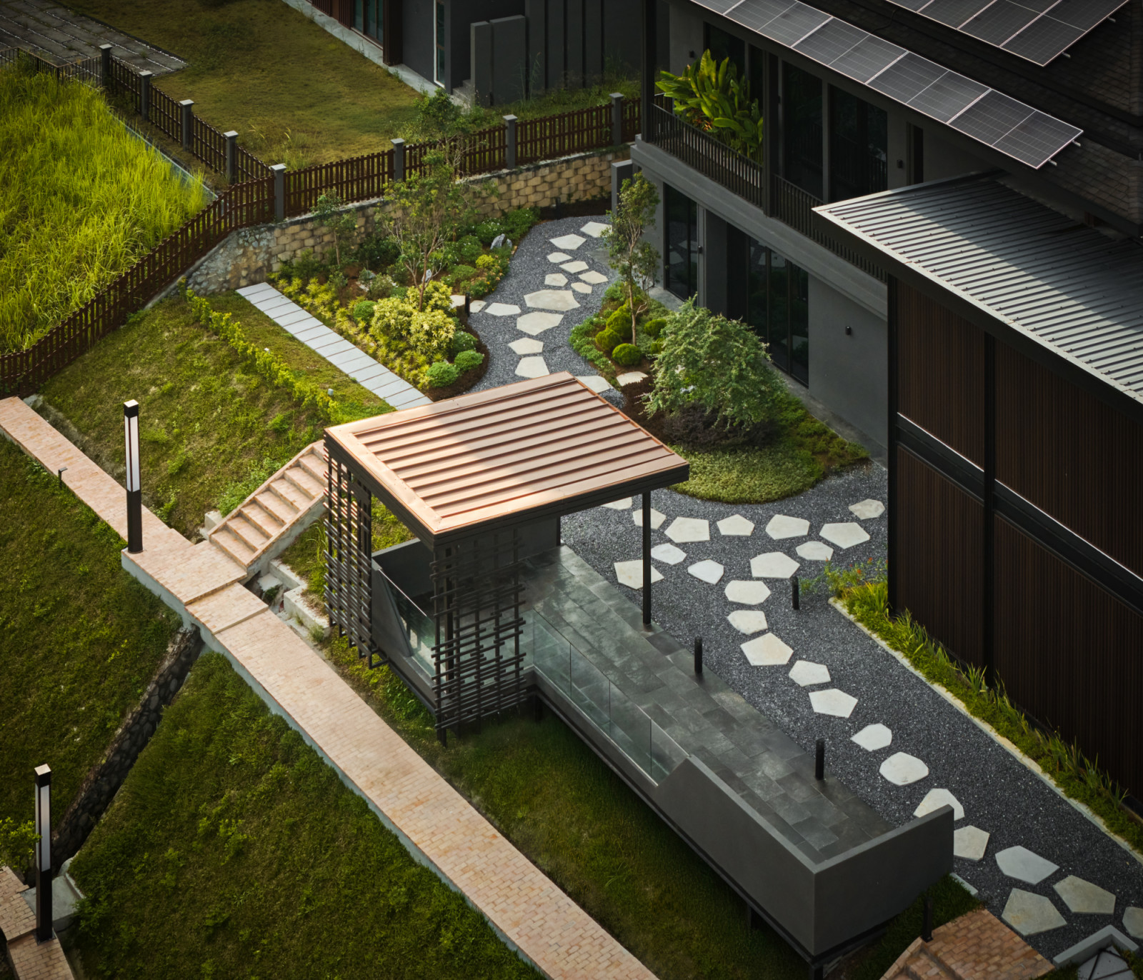 An overhead view of the spring garden shows the irregular white stone pavers set in dark gravel, a timber-roofed pavilion structure, and curated plantings curving toward the house