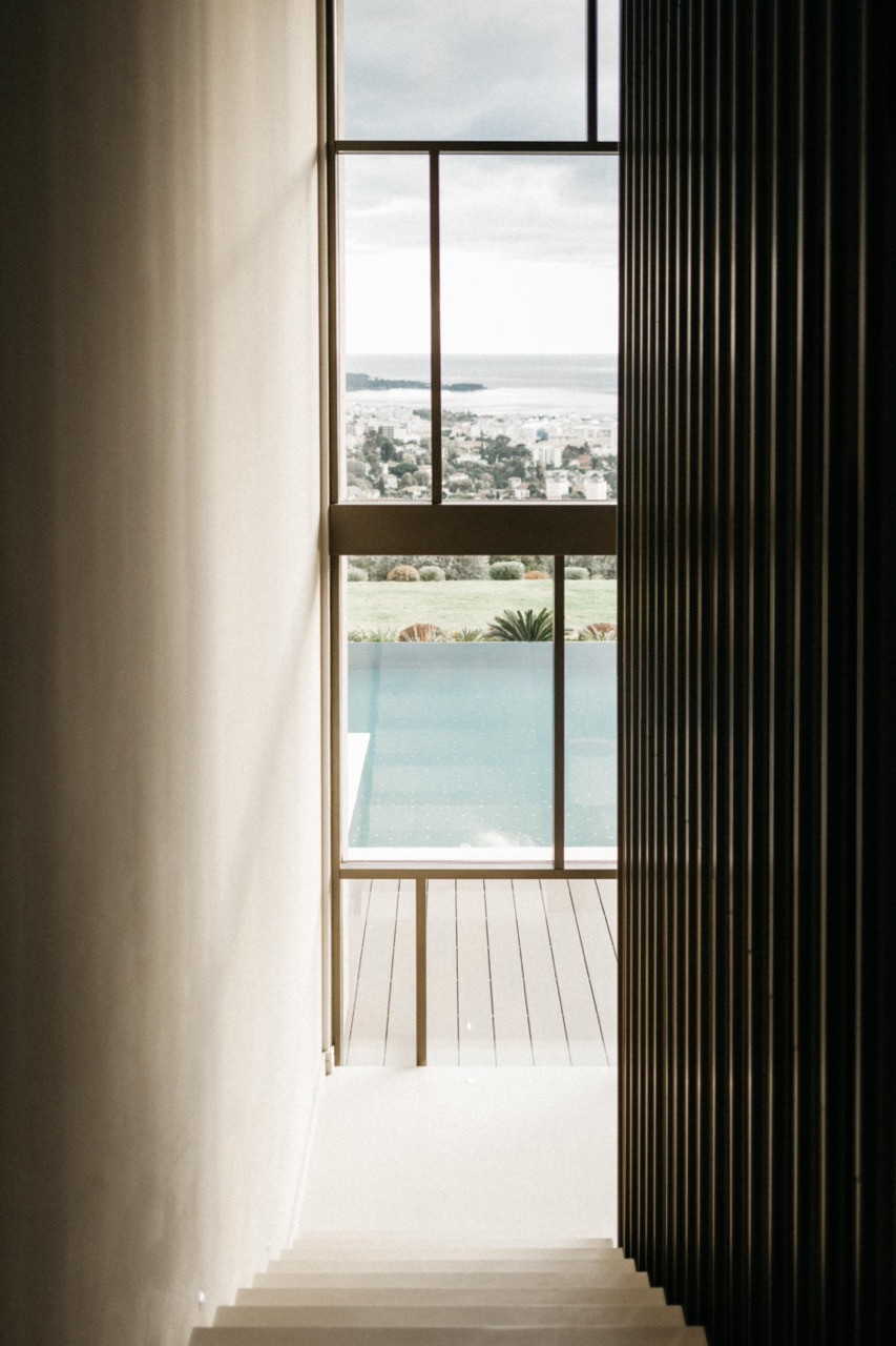 The staircase landing with a full-height steel-framed window overlooking the pool, garden and Cannes below, flanked by a dark vertical timber louvred screen
