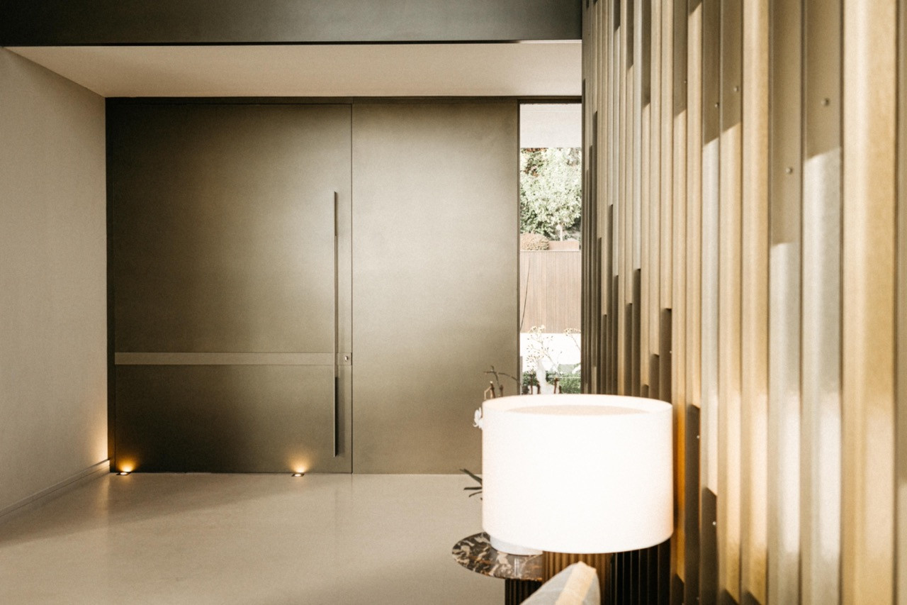 The entrance hall with a large dark steel pivot door, floor-level uplighting, vertical timber louvred wall panel and a cylindrical table lamp in the foreground