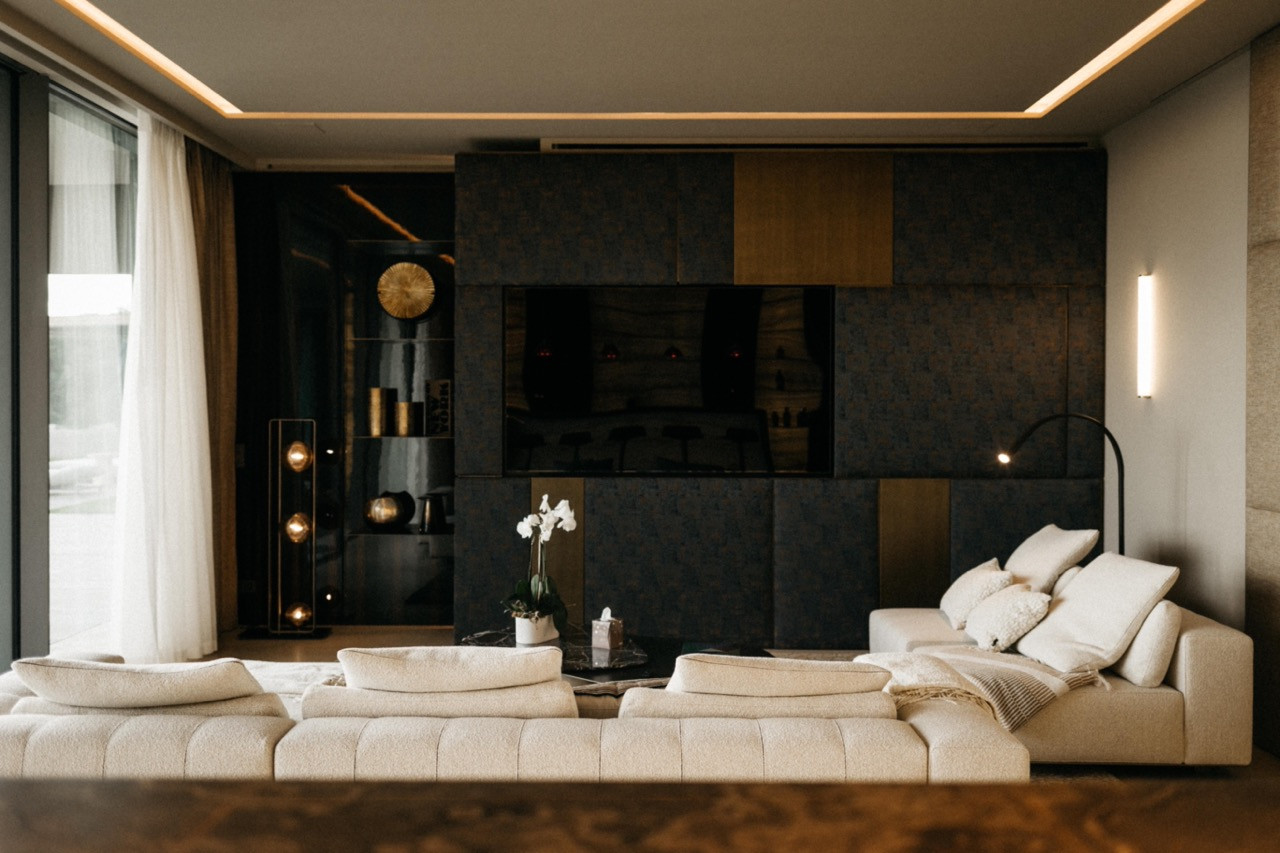 The living room with cream bouclé sofas arranged before a dark textured feature wall with integrated television and gold panel accents