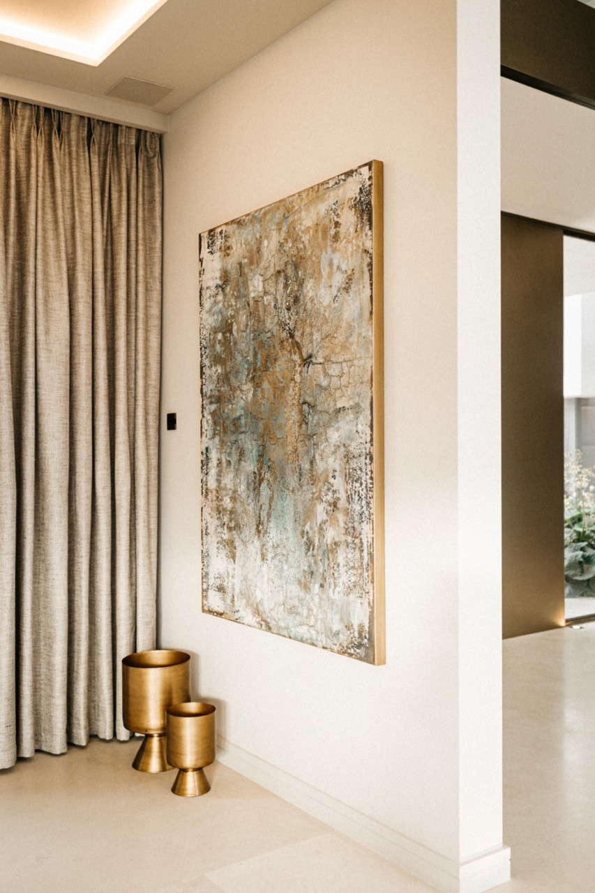 A large-scale abstract painting in gold, teal and white tones on the entrance hall wall, with two brass urn-shaped vessels on the floor below