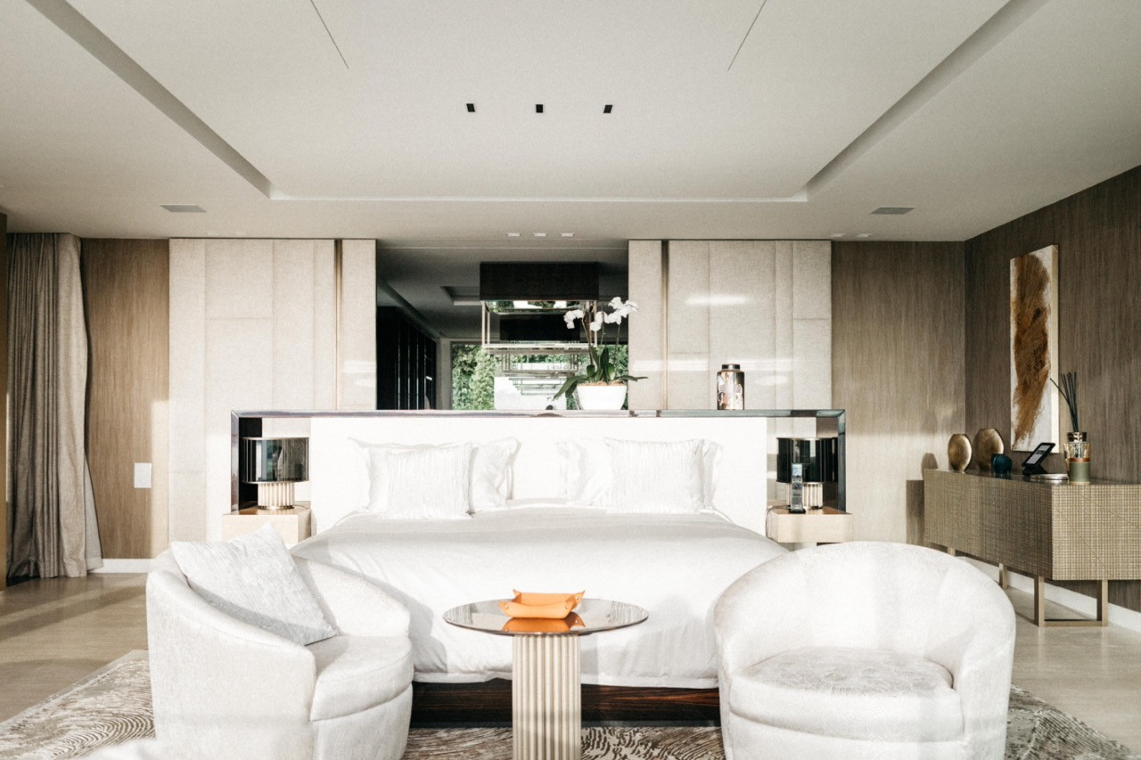 The principal bedroom suite with upholstered headboard wall, mirrored shelf, white armchairs and a fluted column side table