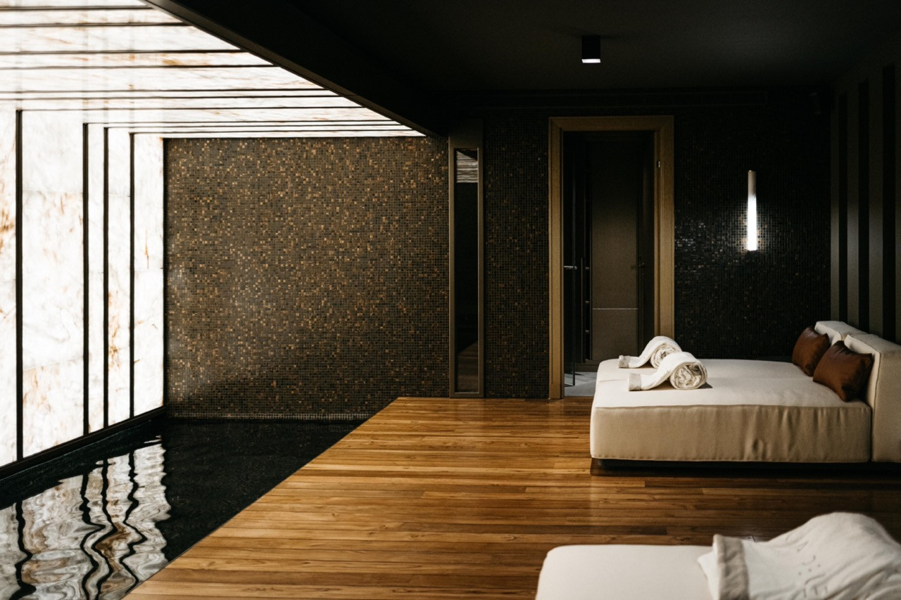 The spa relaxation area features marine teak flooring, dark mosaic walls and a backlit translucent stone wall alongside white upholstered daybeds