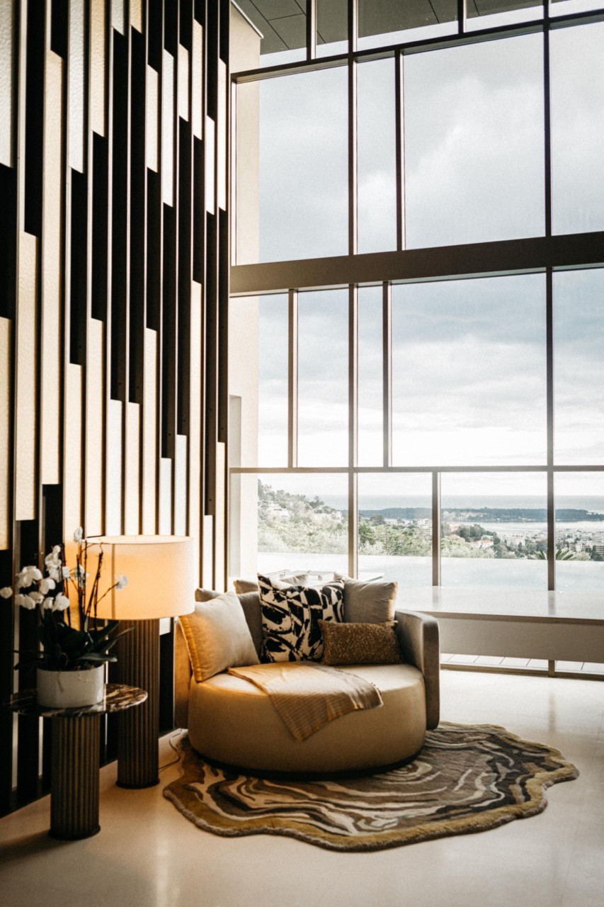 The entrance hall with a double-height steel-framed glazed facade, a round upholstered armchair, agate-patterned rug and vertical metal louvred wall panel