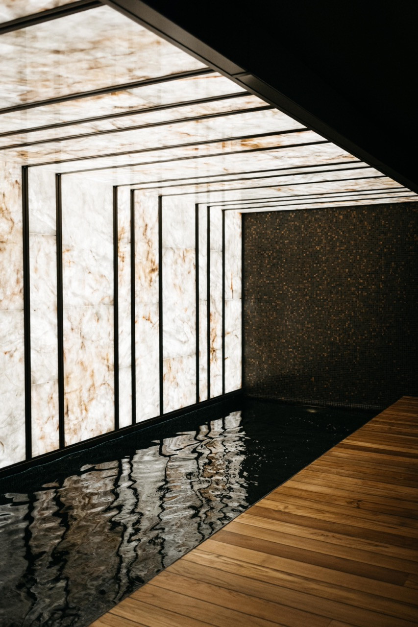 The backlit translucent stone wall, framed in black metal, reflects in the still water of the spa pool