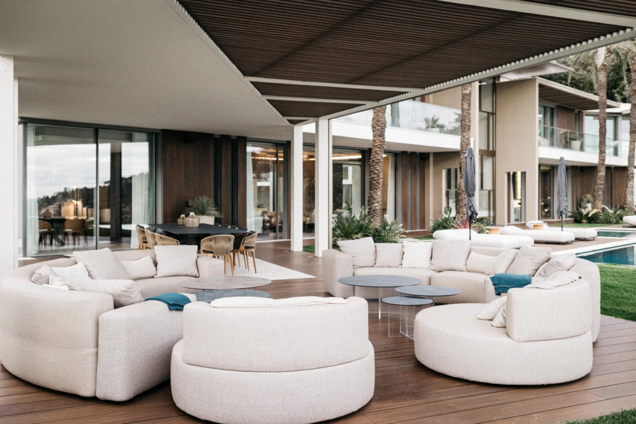 The covered outdoor terrace with circular cream upholstered seating, a teak deck, an outdoor dining area and a pool beyond