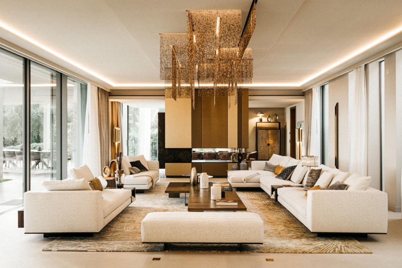 The main living room with cream sofas, walnut coffee table, gold abstract patterned rug and suspended gold mosaic ceiling installation, with floor-to-ceiling glazing overlooking the terrace
